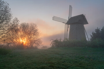 View of an ancient wooden windmill in the Mikhailovskoye estate on a foggy summer morning against the background of the rising sun, Pushkinskiye Gory, Pskov region, Russia