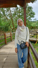 Obraz premium Young Asian woman in hijab and white sweater posing on wooden bridge at park. Fashion lifestyle in nature Indonesia. Beautiful girl with glasses. Sharp focus. Designer.