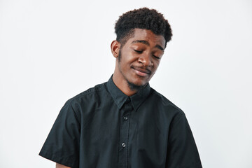 Obraz premium Young African American man with dark skin and curly hair wearing black shirt standing with eyes closed and slight smile on white background in studio portrait.