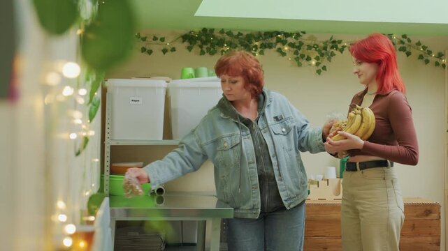 white women exchange bananas in cozy pantry older volunteer in denim jacket offers bananas to younger shopper with red hair, shelves stocked with jars and containers, warm string lights and hanging