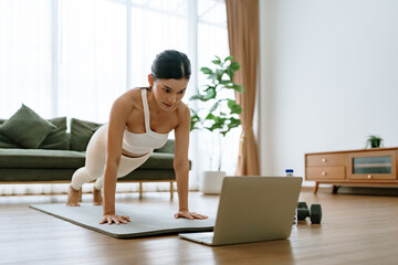 Young Asian woman in sportswear doing plank on training mat while watching online workout tutorials on laptop in the living room. Sport and recreation concept
