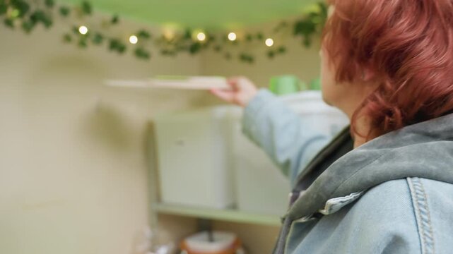 white woman reaches for container, pausing cozy kitchen storage scene with string lights, plastic bins on metal shelf, red hair visible, denim jacket and hoodie, slow deliberate movements, expressions