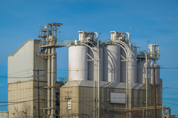 Large industrial concrete plant with tall white silos and metal machinery under a clear blue sky. Wide eye-level shot of heavy machinery and utility lines in daylight.