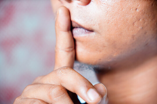 A couple sits on the bed with their backs to each other. A man demonstrates a finger gesture in front of his lips which symbolizes an instruction to be quiet or keep a secret.