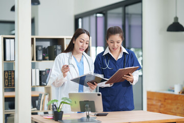Obraz premium Two female healthcare workers, one in blue scrubs and another in a white lab coat, are collaborating and reviewing patient charts on a clipboard and tablet. They are engaged in a professional discussi