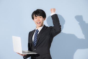 水色背景でパソコンを持つスーツの若い大学生のような男性　新卒や就職のビジネスイメージ　就活の採用イメージカメラ目線　ガッツポーズ	
