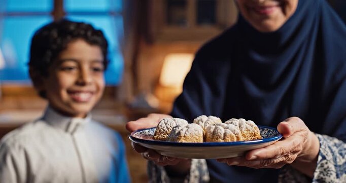 An older woman in a hijab offers a plate of traditional powdered cookies to a smiling young boy, celebrating a festive occasion at home.
