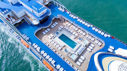 Naklejka premium a luxury cruise ship's open deck featuring a swimming pool, jacuzzis, and rows of sunbeds. Multi-level recreational area with wooden flooring and blue decks over the ocean.