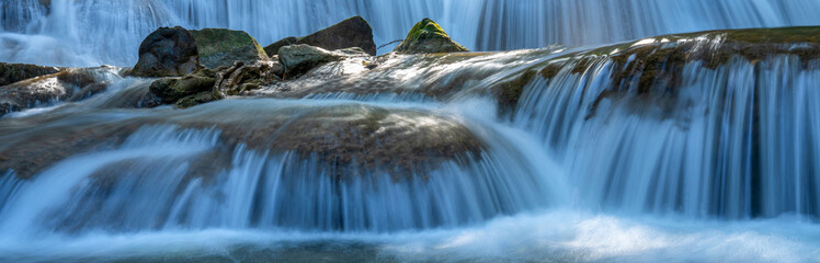 Stunning panoramic close-up of a tiered limestone waterfall with silky water flow. Wide horizontal nature banner background with cascading stream and mossy rocks in a tropical forest. © Yellow Boat