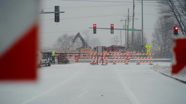 snow-covered intersection is blocked by construction barriers and warning signs while heavy machinery works in the background. winter road construction, traffic control
