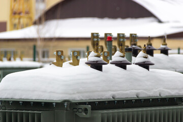 High voltage transformer contacts covered in snow against the backdrop of a house and structure