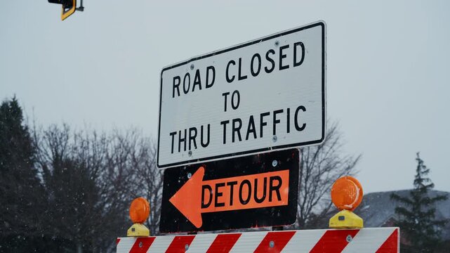 A road closed sign with a detour notice stands on a snow-covered street. traffic control, road safety, transportation disruption caused by snowfall. Close up POV view