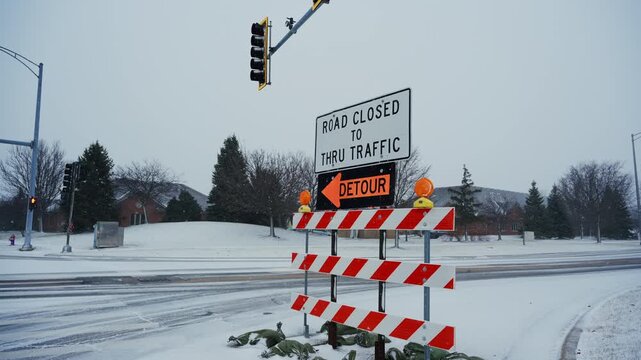 A road closed sign with a detour notice stands on a snow-covered street. traffic control, road safety, transportation disruption caused by snowfall. Close POV view
