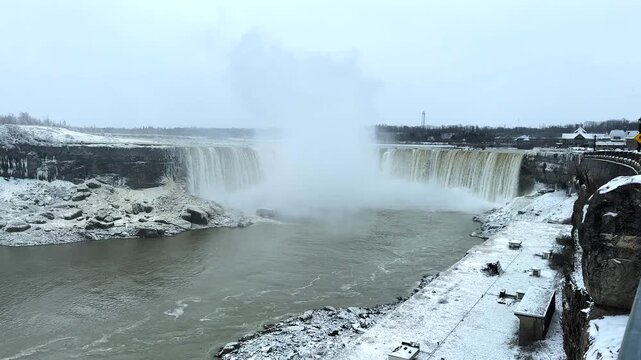 Niagara Falls on a snowy winter day, surrounded by white landscapes as slightly muddy water crashes down, creating a dramatic mist cloud at the base of the waterfall.