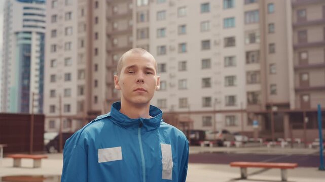 street footballer on wet urban court walking past benches and apartment towers, direct stare to camera, tiny raindrops on hood, assertive posture and steady stride, editorial color tones, close