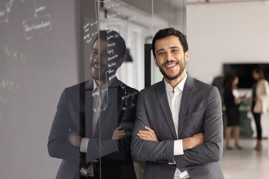 Arabic man, project manager stands confidently with arms crossed leaning against glass wall covered with handwritten business notes and mind maps, smile at camera, exuding professionalism and success