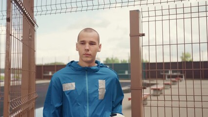 young man holding soccer ball behind rusted fence, urban rooftop court, aspiring striker training alone, blue rain jacket, concrete goals, metal gate, closeup and wide shots emphasizing focused