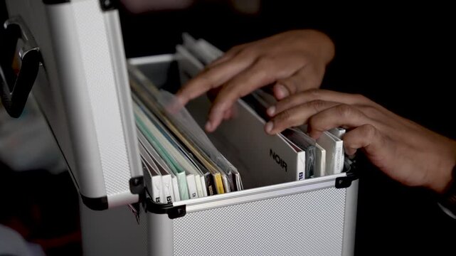A person's hands are shown sorting through vinyl records inside a silver storage box. represents retro music collection collector lifestyle and entertainment concepts against a dark background