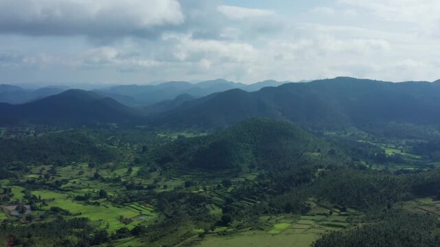 Expansive aerial of Daringbadi's undulating green hills filled with terraced paddy valleys, forested slopes, and distant hazy blue mountains under scattered clouds Odisha, India.