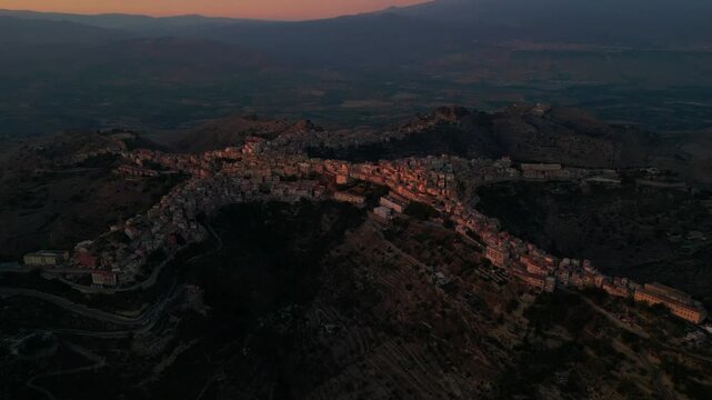Aerial drone footage of Centuripe village at sunset with Mount Etna rising on the horizon. Cinematic drone view over Centuripe revealing its unique hilltop shape beneath the silhouette of Mount Etn