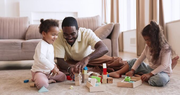 Black father playing developmental games with two little biracial children, build blocks together sitting on floor in living room. Learning through games at home, quality family time and fatherhood