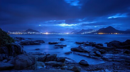 Night Seascape with Rocks and Distant City Lights Under a Deep Blue Cloudy Sky Calm Ocean Water Reflecting Lights Shoreline Rocks in Foreground Long Exposure