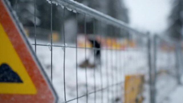 A prominent triangular yellow warning sign, indicating 'work in progress,' stands in a snowy, wintry environment. The sign, with its vivid red border and a black silhouette of a worker with a shovel, 