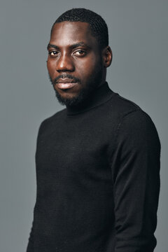 African man portrait in creative conceptual visual style wearing black turtleneck sweater against gray background. Confident young male model with short hair and beard looking at camera.