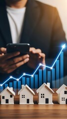 Person in suit using smartphone with rising graph and model houses on table