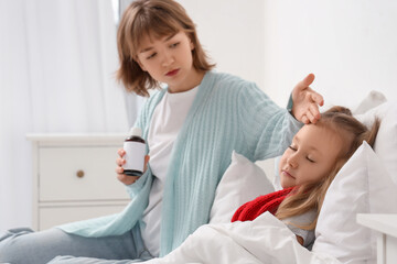 Fototapeta premium Mother with cough syrup checking her ill little daughter's temperature in bedroom