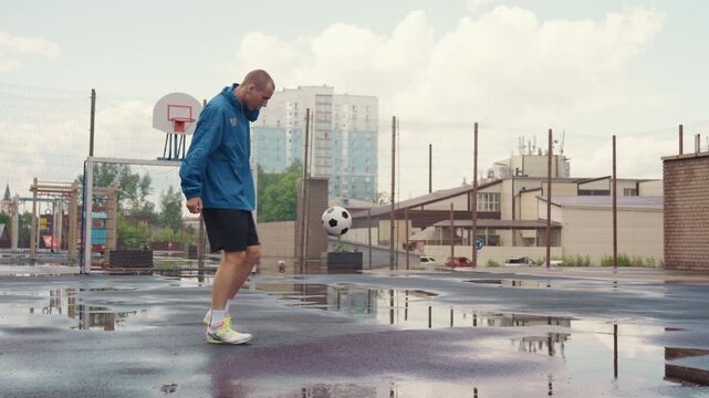 solo football practice on wet court, puddles reflecting skyline, adult male in blue jacket juggling ball near basketball hoop, empty urban lot under overcast sky, steady dribble and controlled pass,