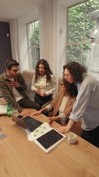 Multicultural team gathers around a laptop at a wooden table, reviewing workflow and ideas, exchanging opinions and focusing on cooperation inside a modern professional office.