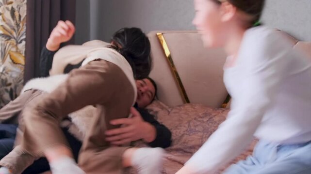 asian father wrestling with children on bed, playful tickles and tumbling, living room turned play zone, laughter and affectionate chaos, relaxed weekend bonding