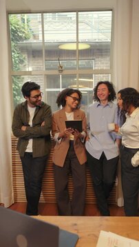 Diverse business people stand by a large window in a modern office, talking openly and sharing ideas while building trust, collaboration, and strong professional connections.