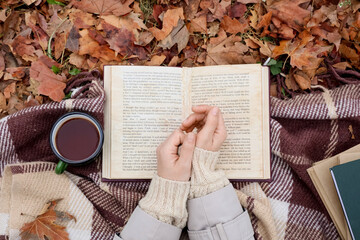 Female hands with book and cup of tea on plaid in autumn park, top view
