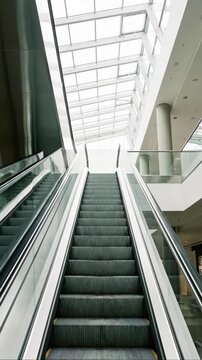 Low angle view of an escalator moving upward in a modern shopping mall with a glass ceiling and bright natural light