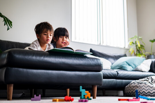 Two young kids sit on a couch, absorbed in a book with colourful blocks scattered nearby
