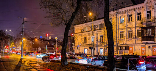 A city at night in a traffic jam. Car lights pierce the tree-lined boulevard. Autumnal evening mood. The way home after work. The architecture old city receding into the distance. Perspective panorama
