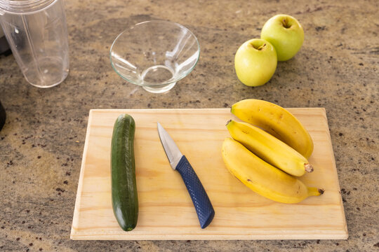 Wooden cutting board is holding cucumber, blue paring knife, bananas on granite counter, copy space
