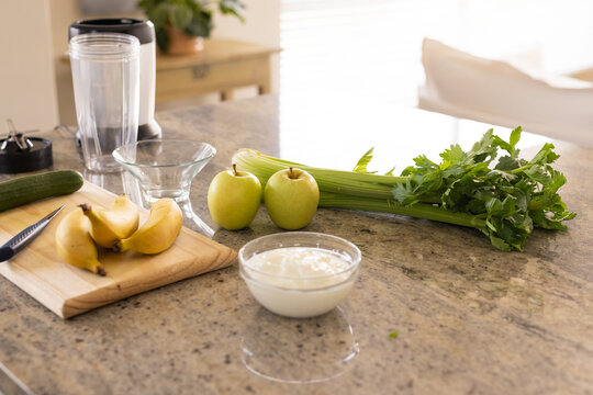 Bananas resting on cutting board with blender, apples, celery and yogurt sitting on granite counter