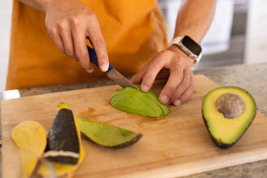 Peeled avocado being sliced on wooden cutting board on speckled countertop near avocado half