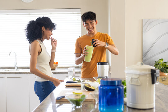Diverse couple in jeans preparing drink at kitchen island using green shaker cup and supplements