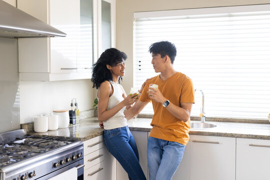 Diverse couple leaning at speckled stone countertop in kitchen, holding clear glasses and talking