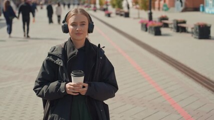 young woman with coffee and headphones pauses on city plaza wearing black jacket and green pants candid portrait shows relaxed expression as she listens to music while commuters pass by, tram tracks