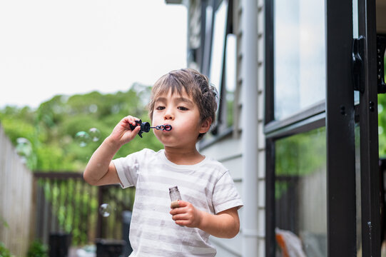 A cheerful boy stands in a backyard, happily blowing bubbles with a wand in hand