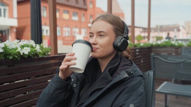 White woman outdoor cafe sipping coffee, wearing black jacket and wireless headphones urban terrace with planters and brick buildings candid closeup shows playful smirk evolving into raised eyebrow