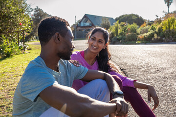 Diverse couple sitting on suburban street, resting after run, checking fitness watch