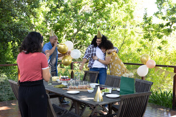 Obraz premium Diverse friends hugging and clapping around table on deck with balloons, pastries, gift bag