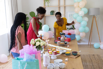 African American friends preparing cupcakes on table under pastel balloons in bright dining room