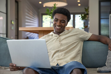 African american man smiling while using laptop on couch in living room with table, potted plants